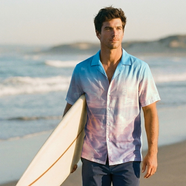 Man walking on a beach holding a surfboard, wearing a patterned shirt and shorts.