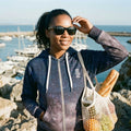 Woman standing by a marina holding a bag of groceries, wearing sunglasses and a hoodie.