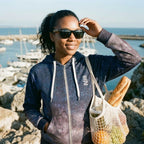 Woman standing by a marina holding a bag of groceries, wearing sunglasses and a hoodie.