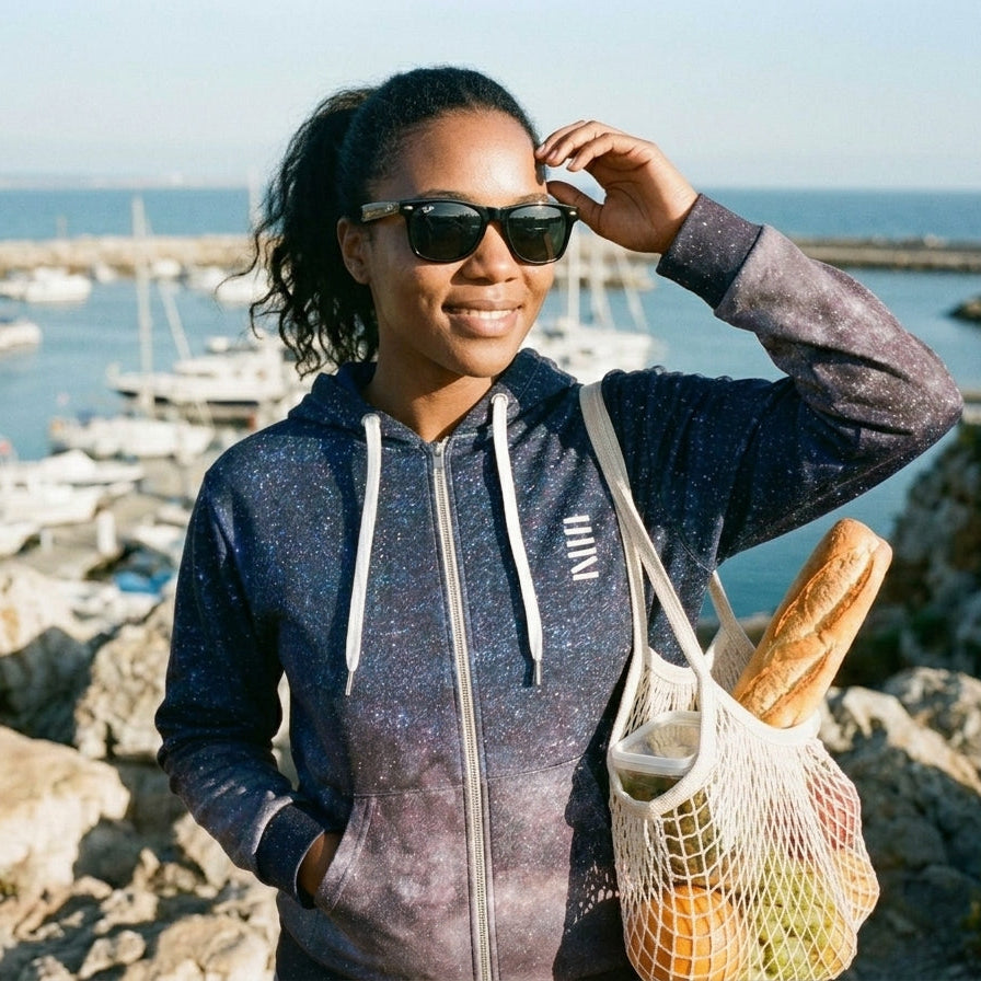 Woman standing by a marina holding a bag of groceries, wearing sunglasses and a hoodie.